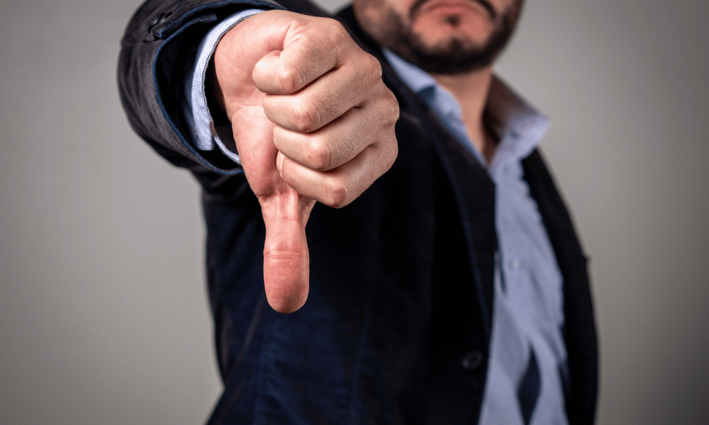 kivitelezés: egy szakállas férfi ingben és elegánsabb kabátban, előtérben az egyik keze lefele mutató hüvelykujjal