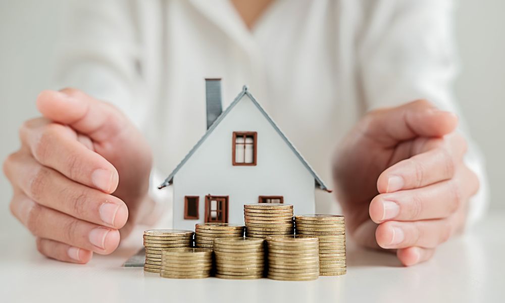 homlokzatszigetelés: előtérben egy kicsi játékház, előtte néhány oszlop fémpénz, a háttérben egy fehér felsős ember, aki a két keze között tartja a kompozíciót