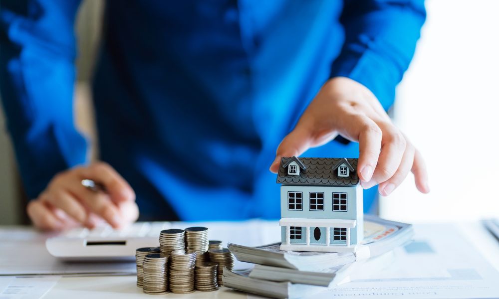 homlokzatszigetelés: előtérben egy kicsi játékház, előtte egy köteg papírpénz és pár oszlop fémpénz, a háttérben egy kék inges férfi, egyik kezével egy számológépet nyomkod, a másikkal a házat rakja le